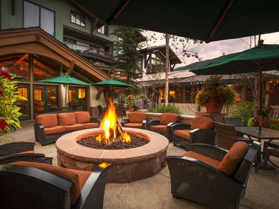 A circular stone fire pit surrounded by wicker chairs and tables, with green umbrellas providing shade.