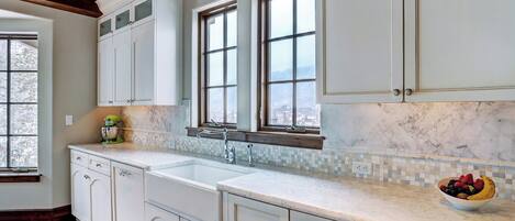 A kitchen with white cabinets, a marble countertop, a window with a view, and a bowl of fruit.