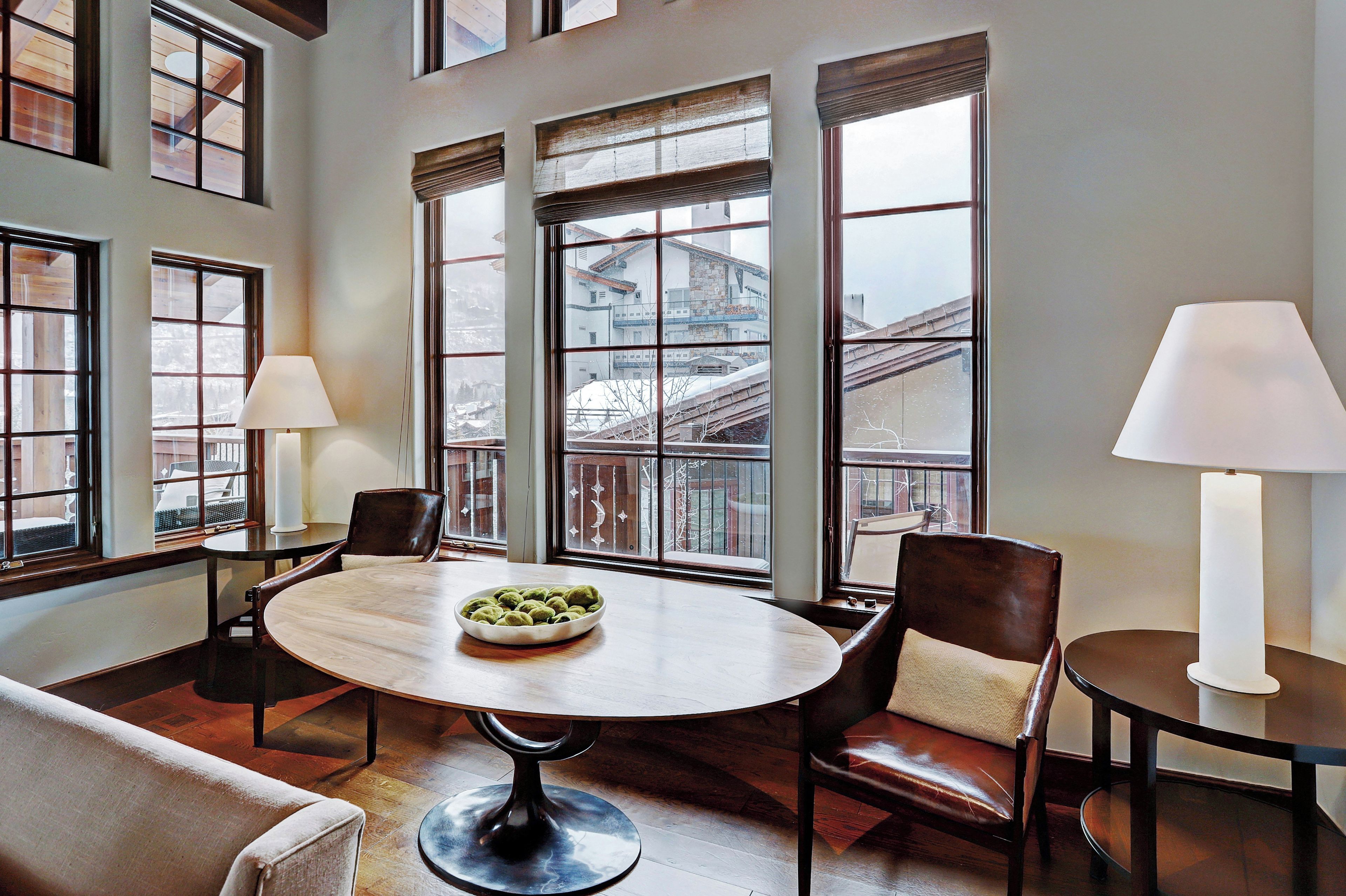 A room with a round table, two chairs, a bowl of snacks, and large windows with a view of buildings.