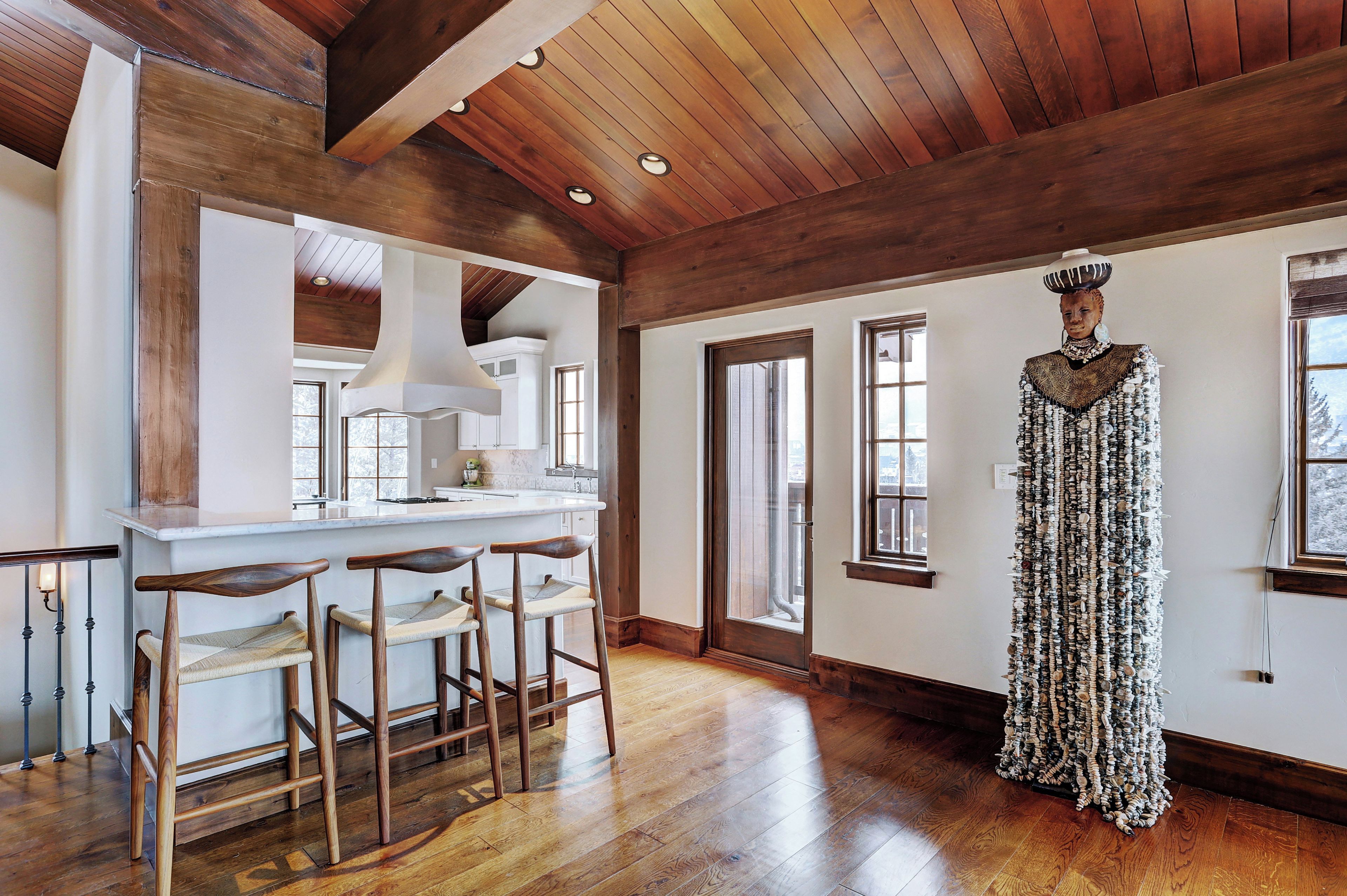 A spacious room with a wooden ceiling, a kitchen area with a white countertop and stools, and a large window.