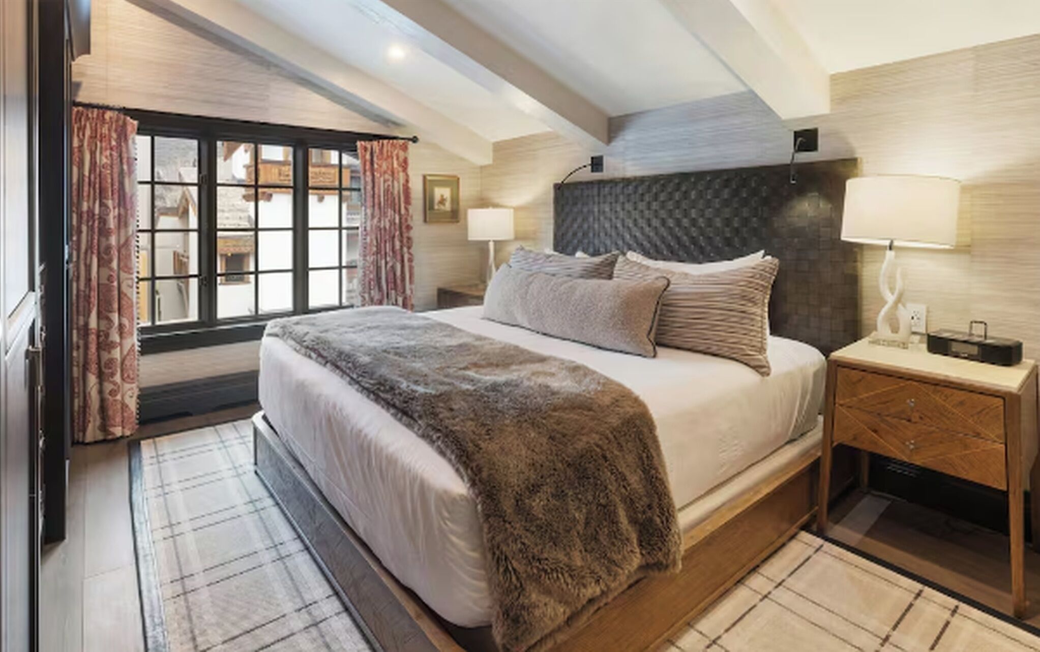 Ein Schlafzimmer mit einem großen Bett, einem Nachttisch, einer Lampe und Blick durch ein mit Vorhängen versehenes Fenster.