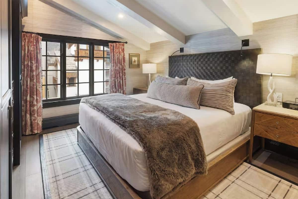 A bedroom with a large bed, a nightstand, a lamp, and a view through a window with curtains.