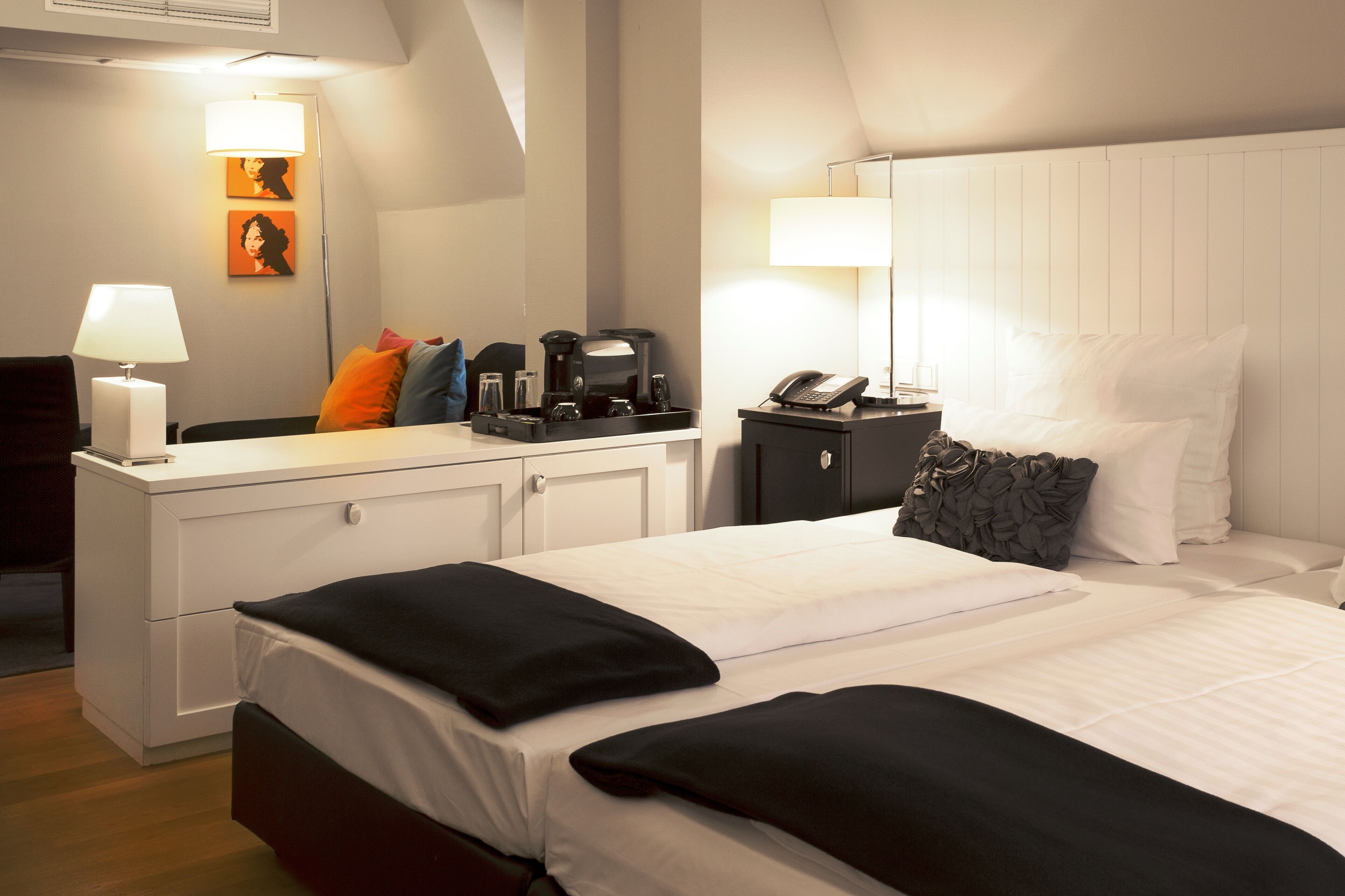 A hotel room with a neatly made bed, a bedside table with a lamp, a coffee maker, and a phone. There is a desk with a lamp and a chair, and artwork on the wall.