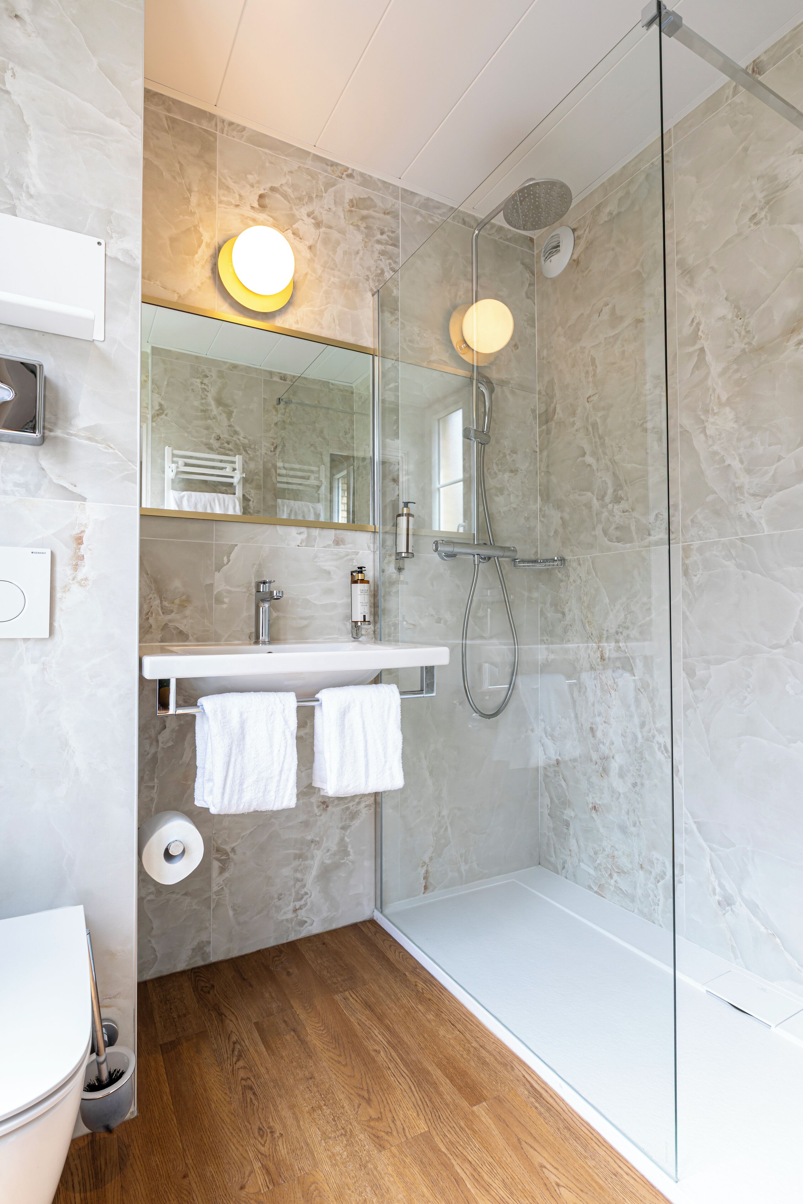 Une salle de bain moderne avec une douche à parois de verre, un lavabo blanc avec un miroir au-dessus et un plancher en bois.