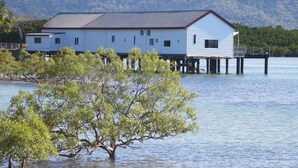 A white building with a dark roof, situated on stilts over water, surrounded by trees.