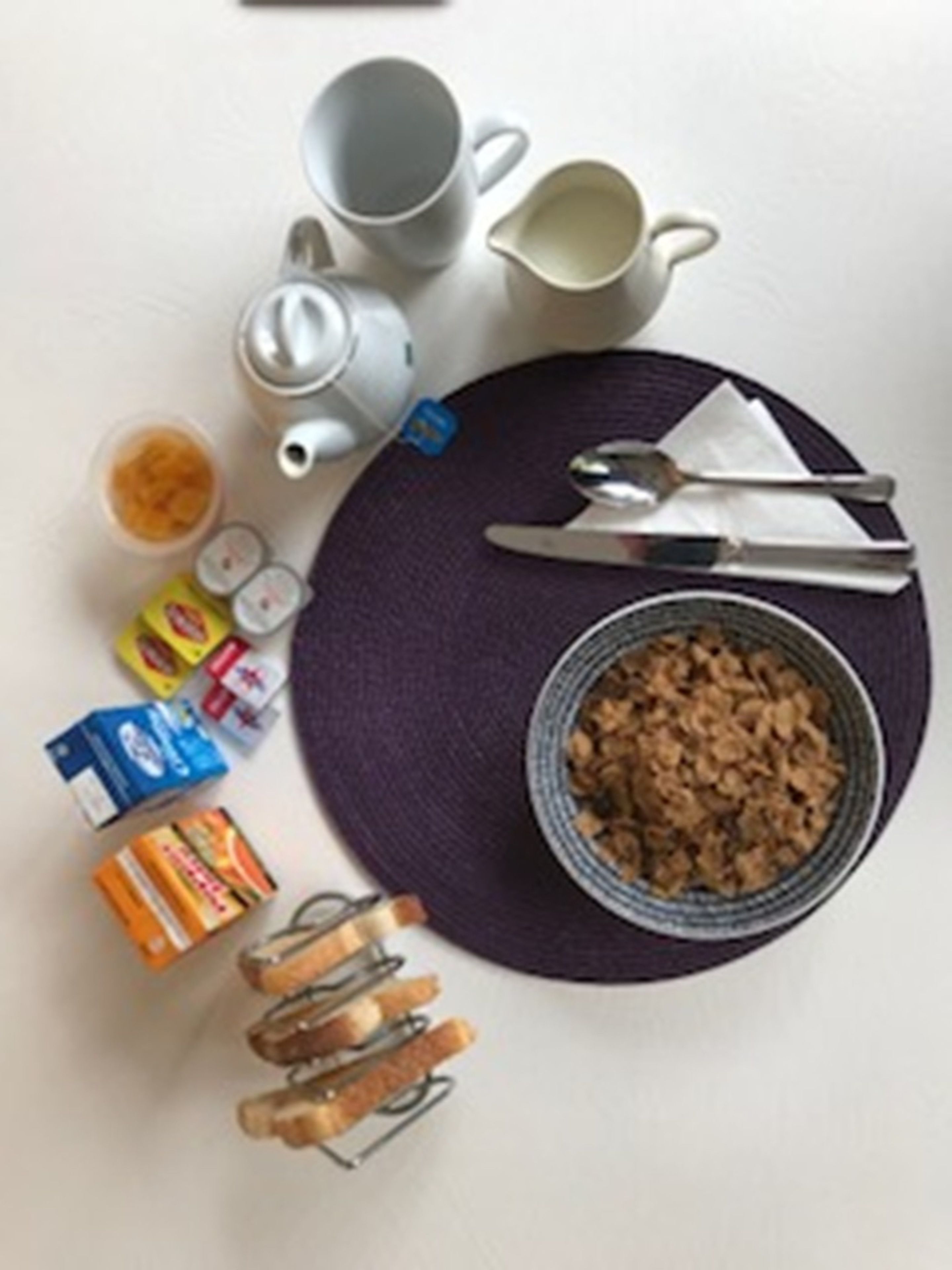 A table with a bowl of cereal, a teapot, a cup, a spoon, a knife, a napkin, and some packaged food items.