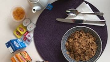 A table with a bowl of cereal, a teapot, a cup, a spoon, a knife, a napkin, and some packaged food items.