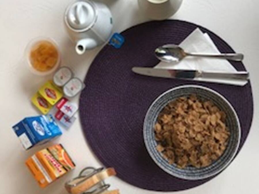 A table with a bowl of cereal, a teapot, a cup, a spoon, a knife, a napkin, and some packaged food items.