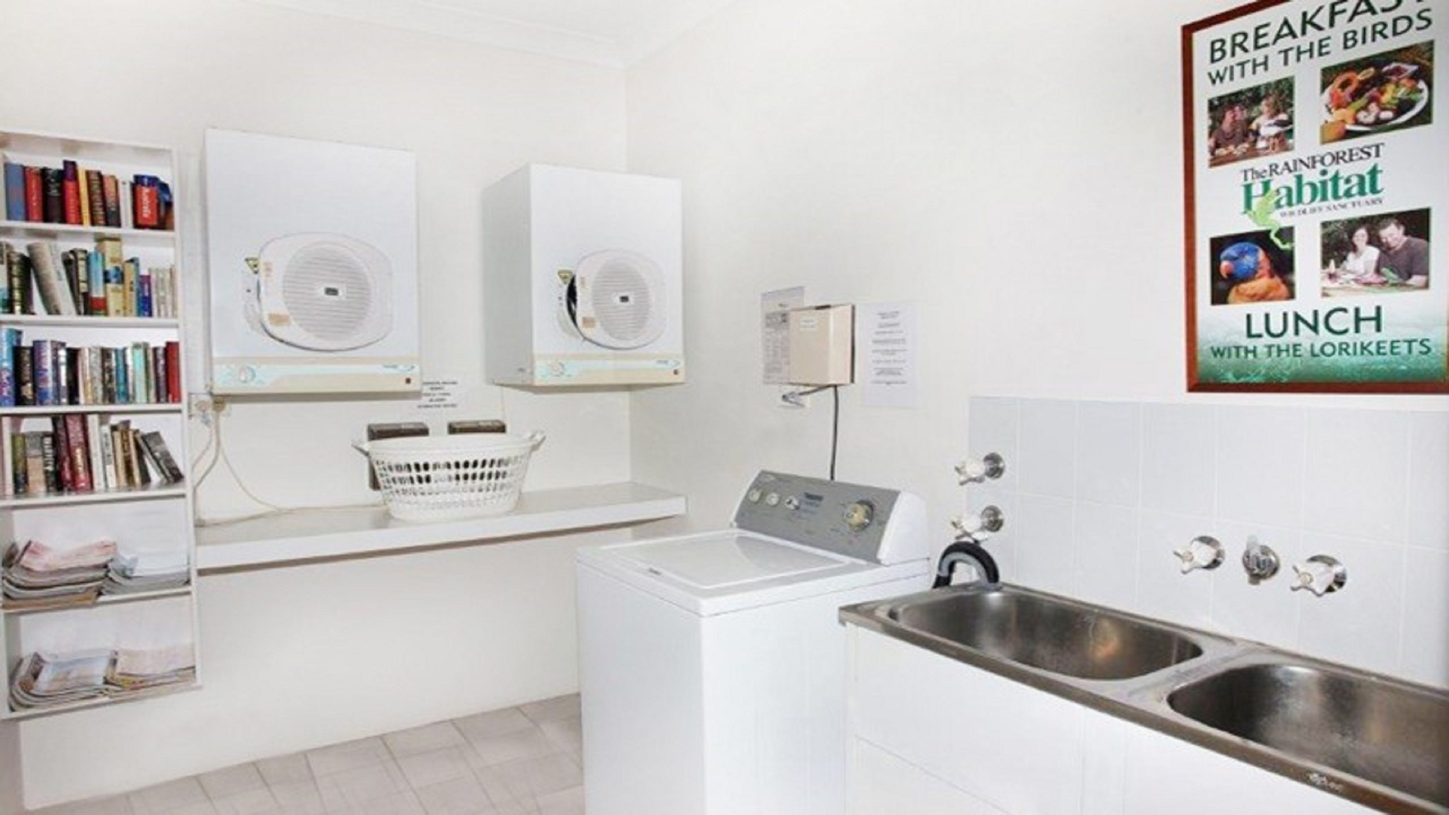 A laundry area with two dryers, a washing machine, and a sink. There is a bookshelf with books and a poster on the wall.