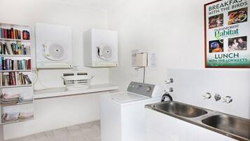 A laundry area with two dryers, a washing machine, and a sink. There is a bookshelf with books and a poster on the wall.