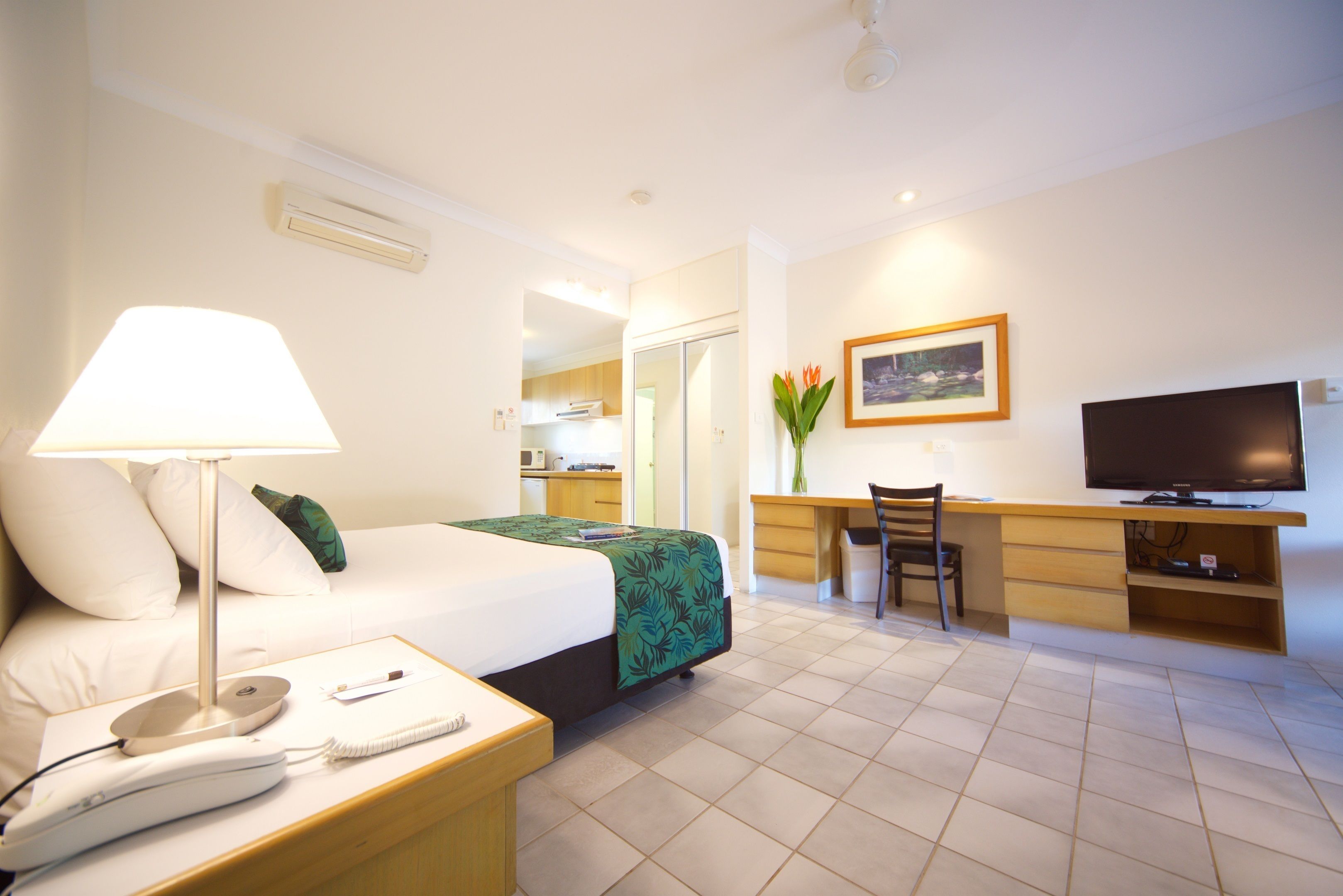 A hotel room with a bed, bedside lamp, desk, chair, television, and a painting on the wall.