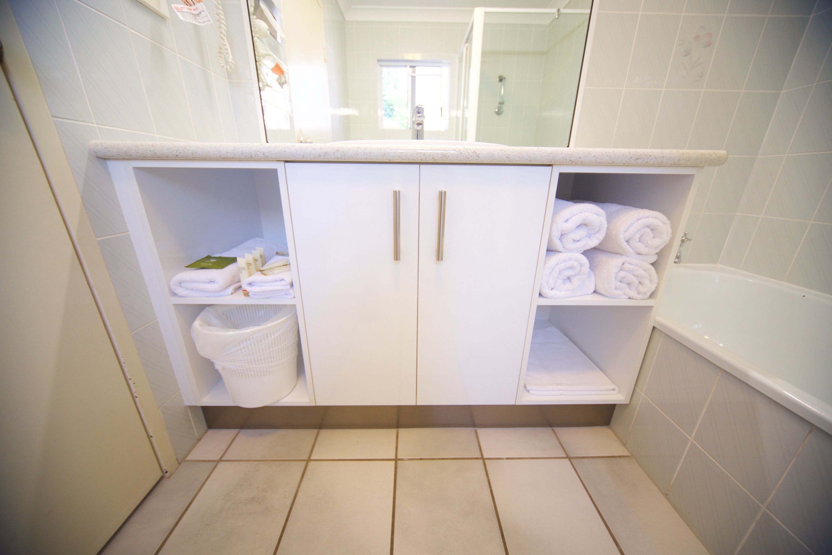 A bathroom with white tiled walls, a white sink with a countertop, two cabinets with handles, a white laundry basket, and a white bathtub.