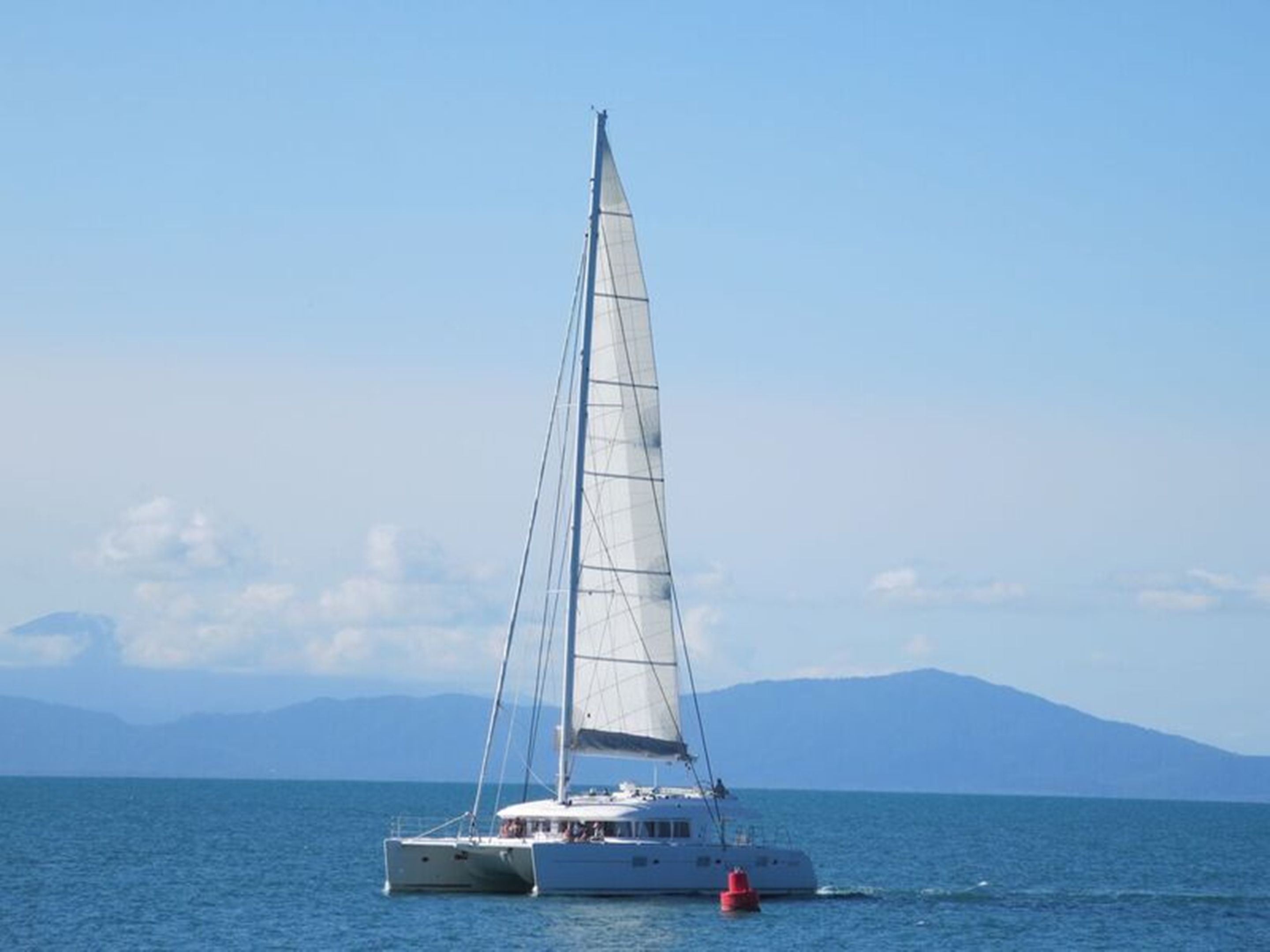Un voilier sur une eau calme, sous un ciel dégagé, avec des montagnes qui se dessinent à l’horizon.