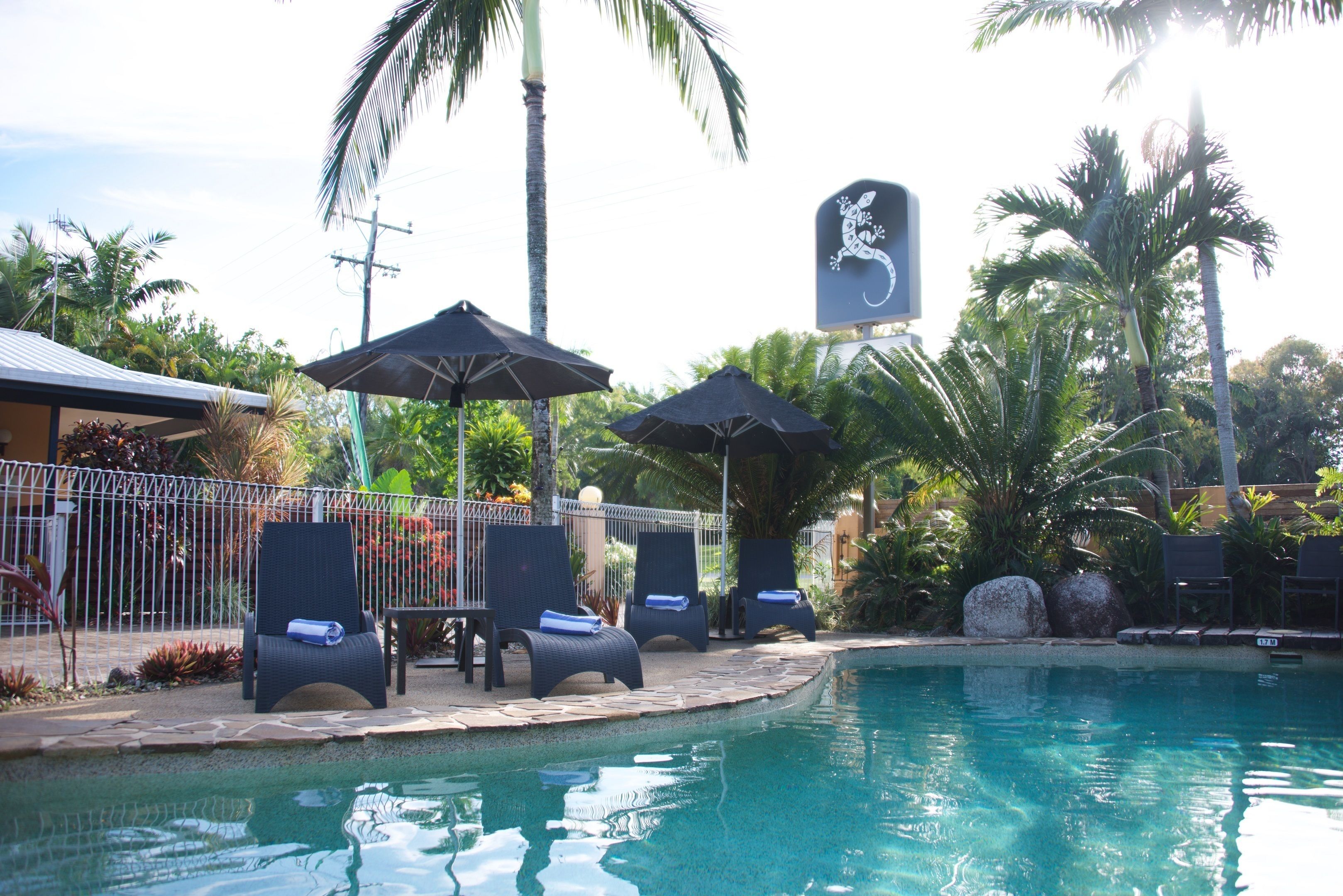 A pool area with lounge chairs and umbrellas, surrounded by palm trees and a fence.