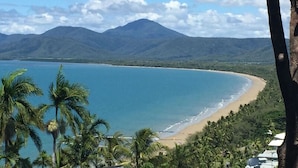 A coastal landscape with a beach, palm trees, and mountains in the background.