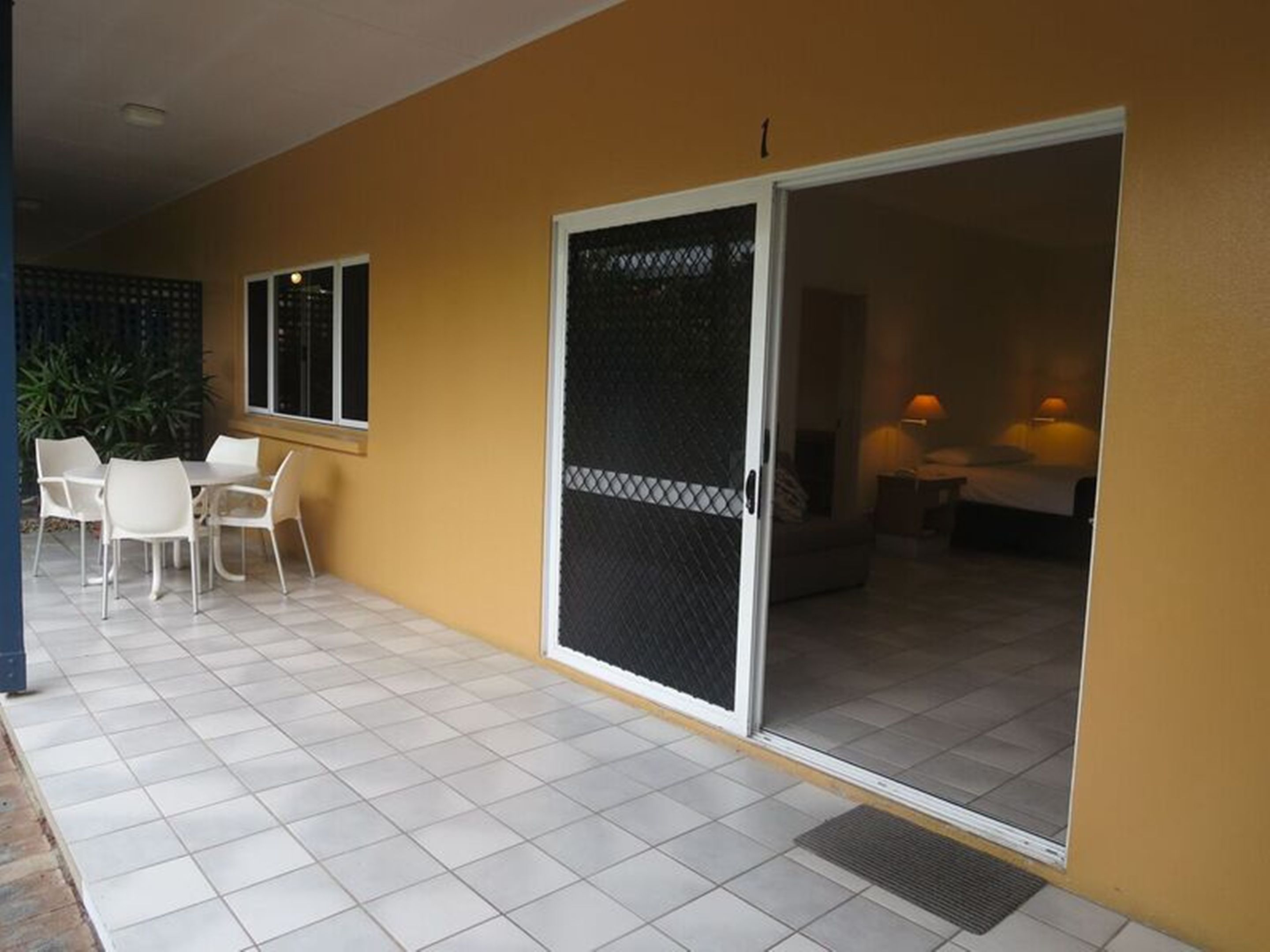 Un patio coperto con pavimento piastrellato bianco, una zona salotto con sedie bianche e una porta scorrevole in vetro che dà su una camera da letto con un letto e un comodino.