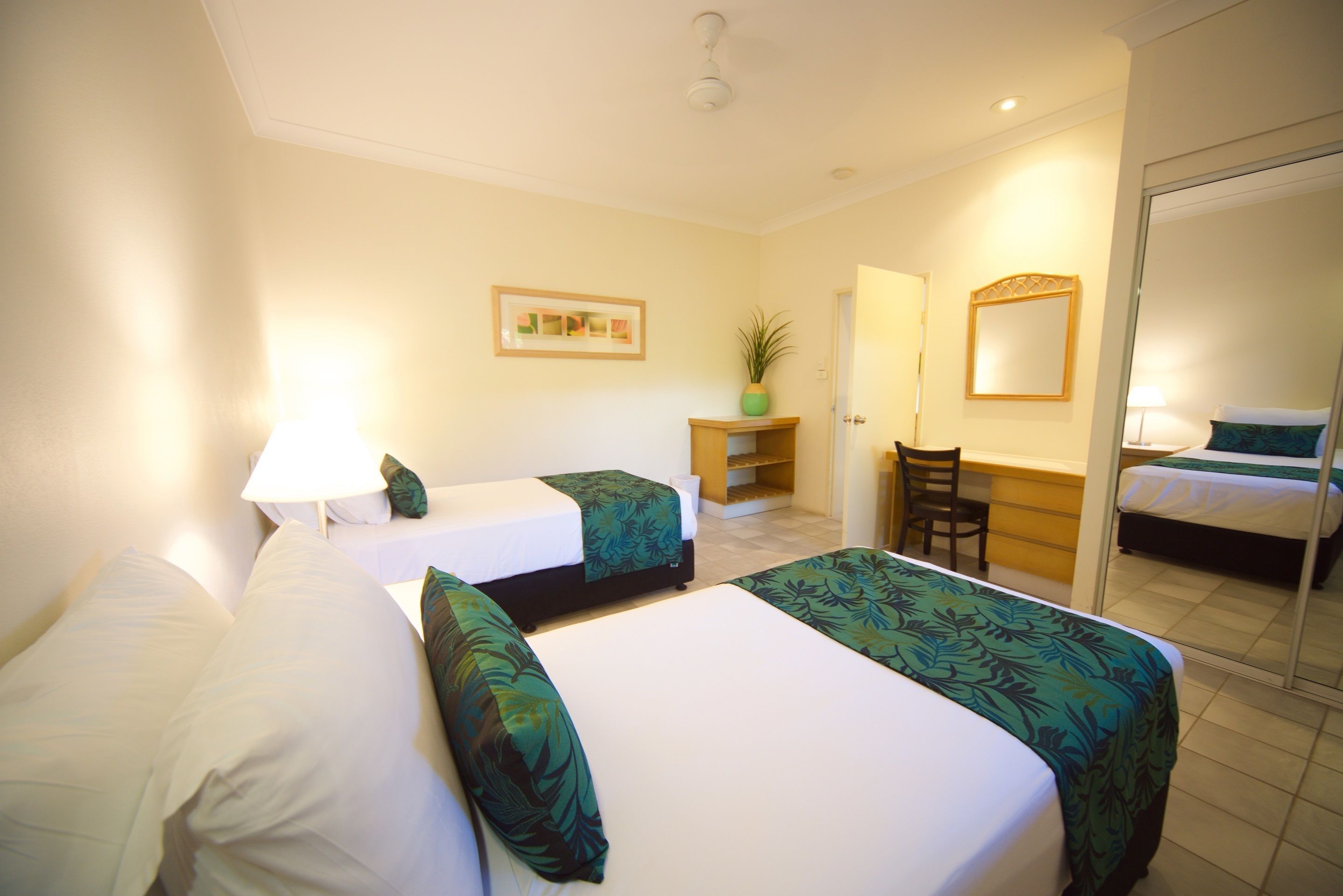 A hotel room with two beds, a desk, a chair, a mirror, and a plant.