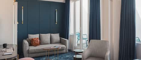 A modern hotel room with a grey sofa, a small round table, a pink ottoman, a grey armchair, and a large bed with a blue headboard.