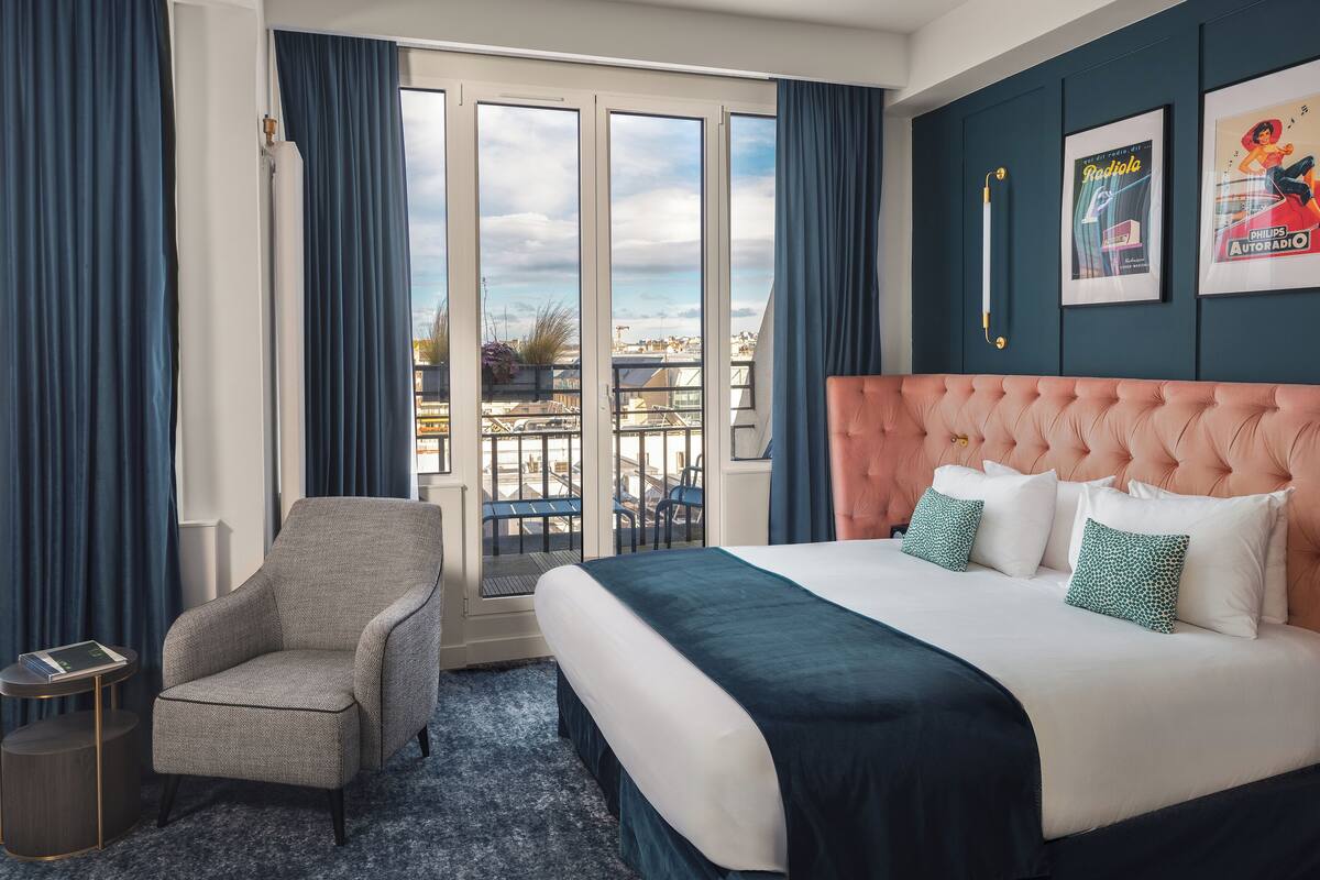 A hotel room with a large bed, a chair, a small table, and a view of the city through large windows.