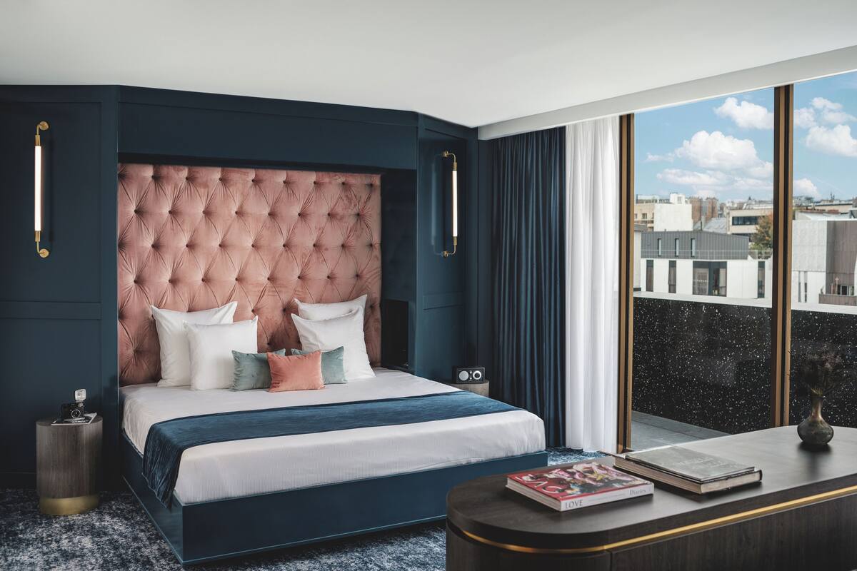 A modern hotel room with a large bed, a nightstand, a desk, and a view of the city through large windows.