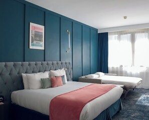 A hotel room with a large bed, a smaller bed, a blue accent wall, a framed picture, and a window with curtains. - Pley Hotel (Paris)