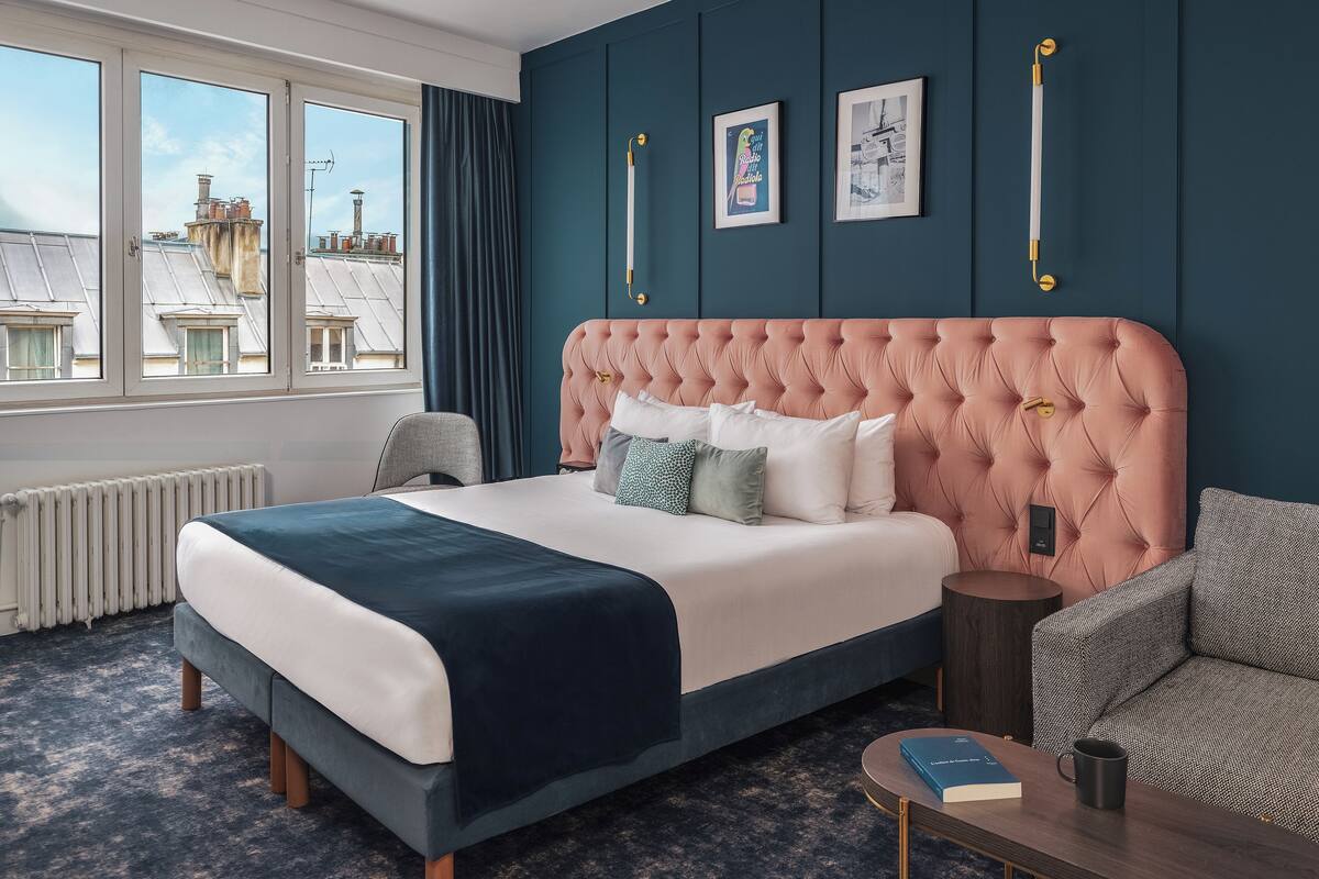 A bedroom with a tufted headboard, a large bed with multiple pillows, a bedside table with a book and a mug, a chair, and a view of buildings through large windows.