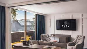 Camera d'albergo moderna con una grande finestra, una TV a schermo piatto, un divano, due poltrone, un tavolino e un minibar.