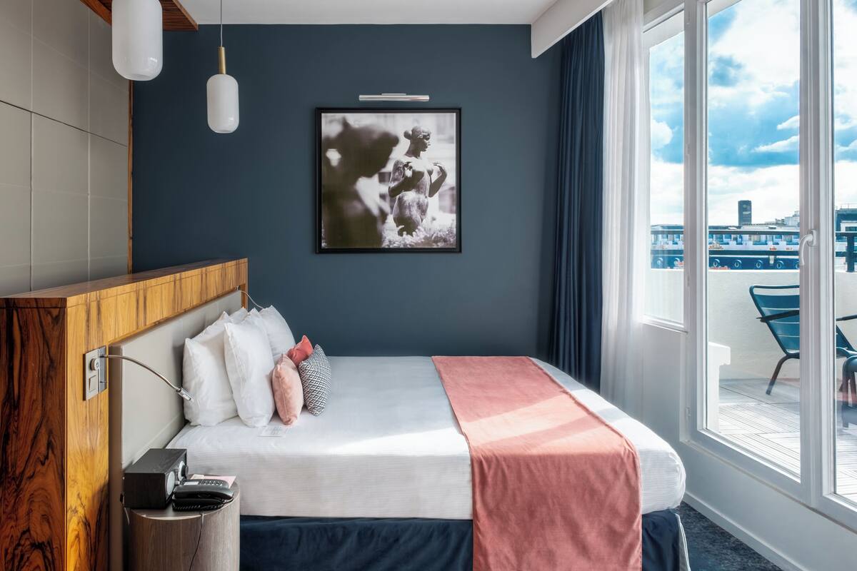 A modern hotel room with a large bed, a wooden headboard, a bedside table with a phone and a trash bin, a framed picture on the wall, and a view of the city through large windows.