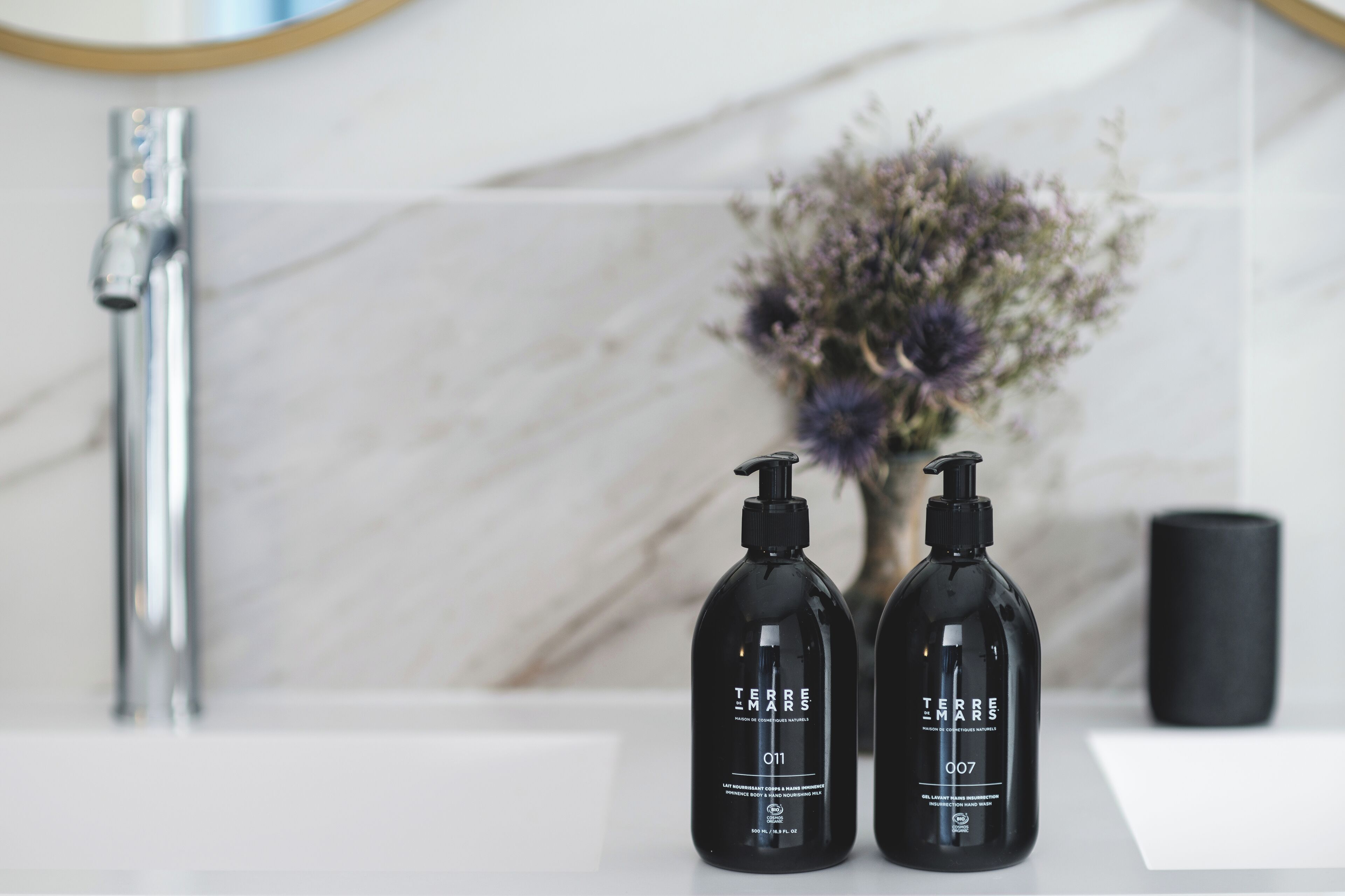 Dos botellas negras de productos para el cuidado de la piel Terre Mars sobre una encimera de baño blanca con salpicadero de mármol y un grifo moderno.
