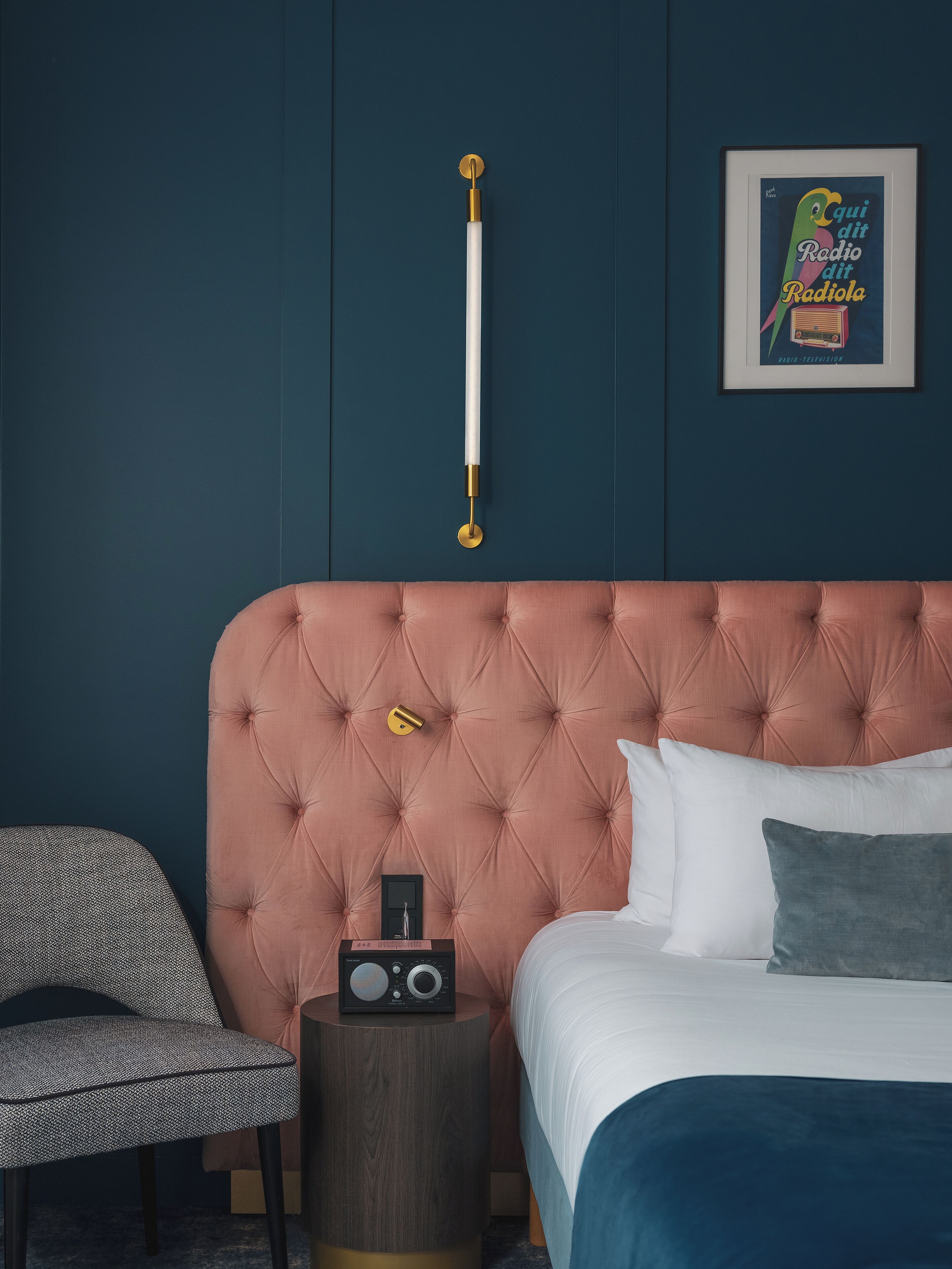 A bedroom with a tufted headboard, a bedside table with a radio, a framed picture on the wall, and a chair.