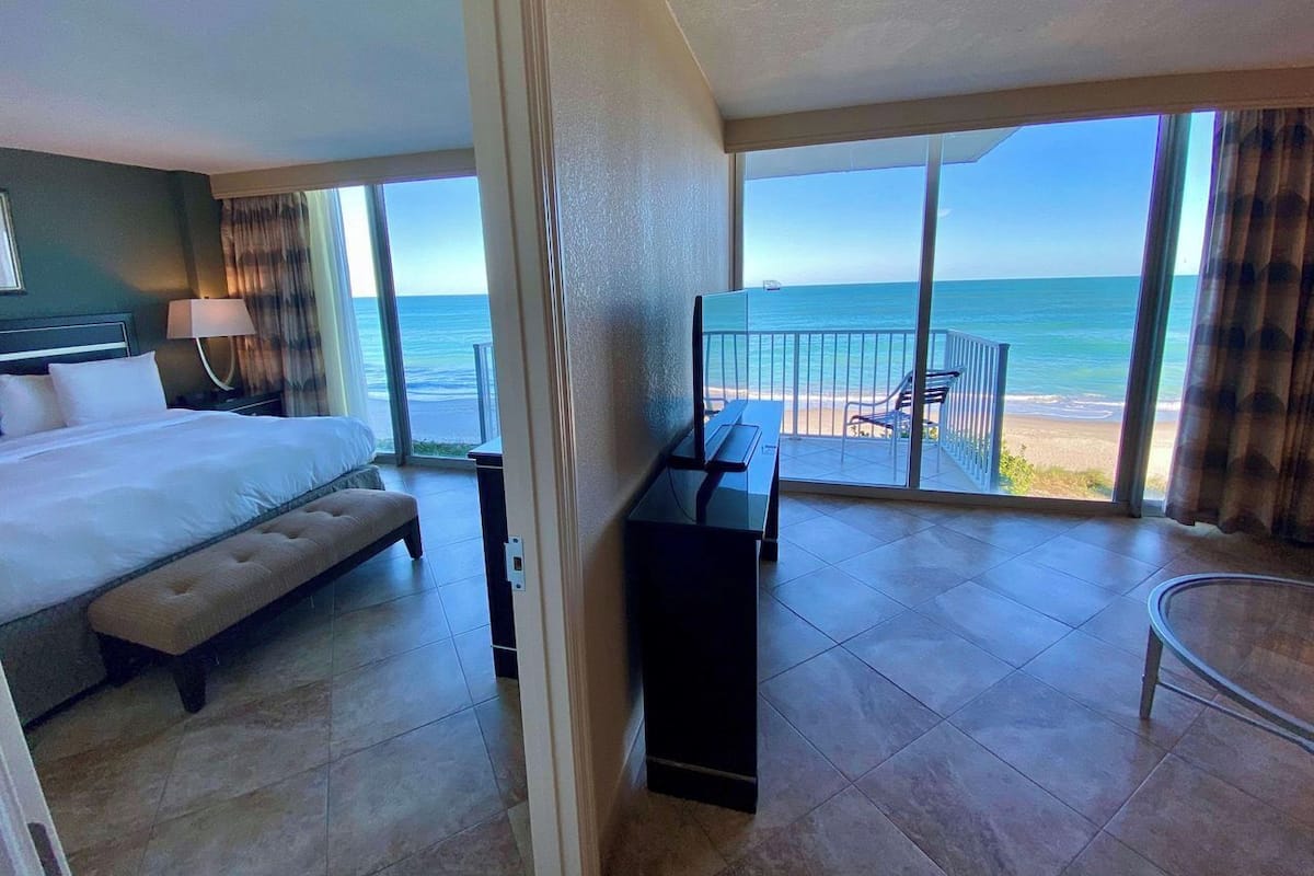A hotel room with a bed, a desk, and a balcony with a view of the beach and ocean.