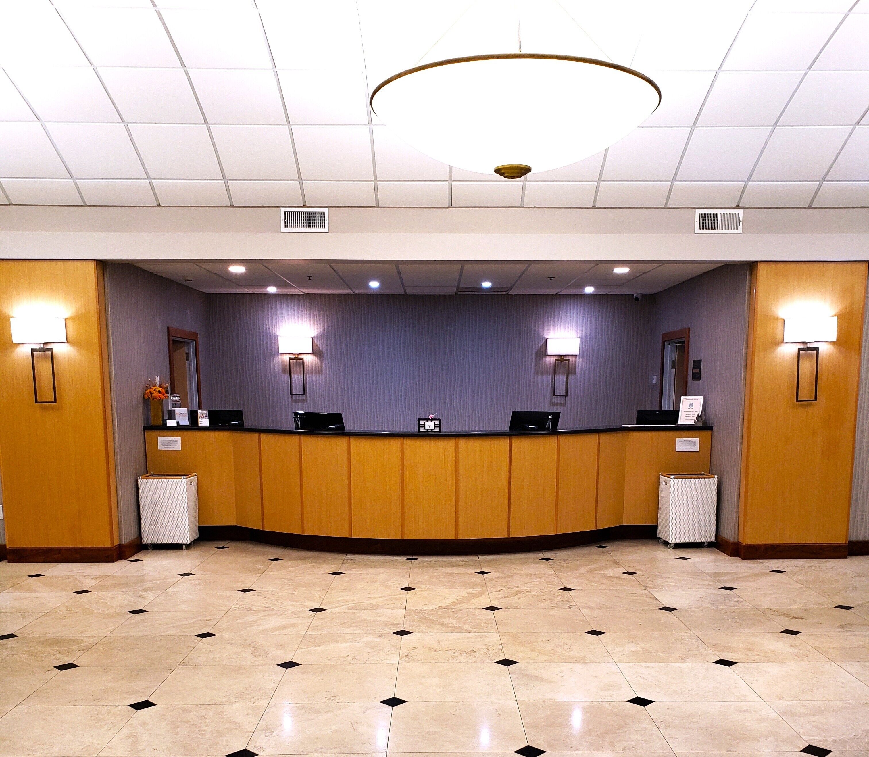 Una zona de recepción con mostrador curvo, dos estaciones de trabajo con computadoras y dos lámparas fijadas a la pared.