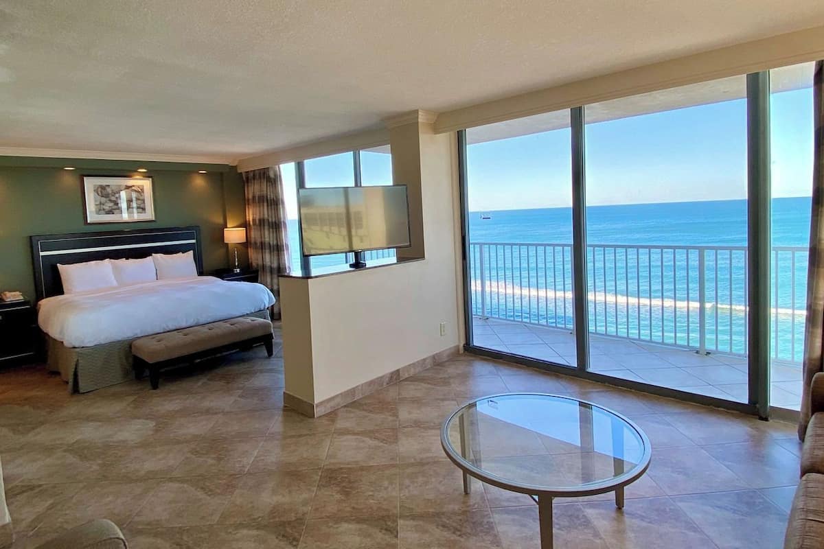 A hotel room with a bed, a glass table, a TV, and a view of the ocean.