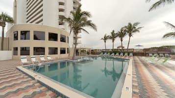 Una piscina con sillones de descanso y palmeras, rodeada por un edificio con balcones.