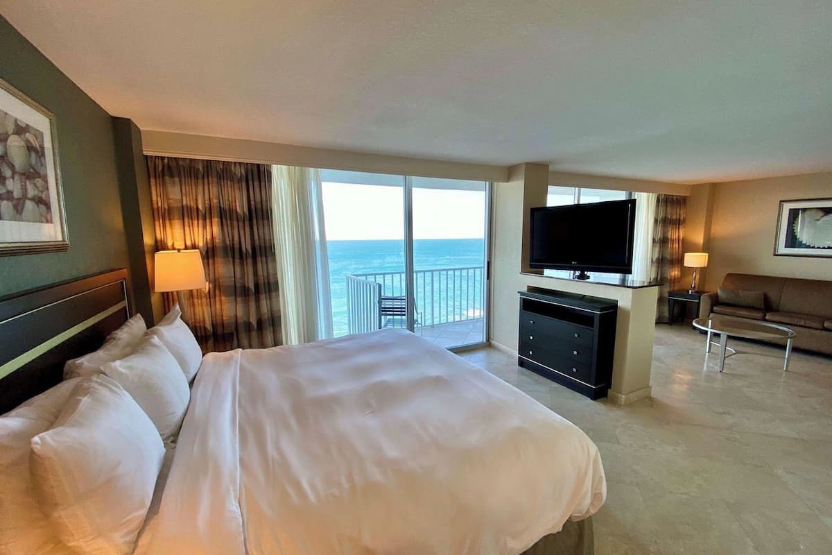 A hotel room with a large bed, a television, a desk, and a balcony with a view of the sea.