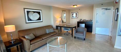 A living room with a brown sofa, a glass coffee table, a gray armchair, and a dining table with chairs. There is a kitchen area with a sink and a refrigerator.