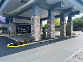 Entrada de un edificio con pilares de piedra, cielo despejado y zona de estacionamiento.