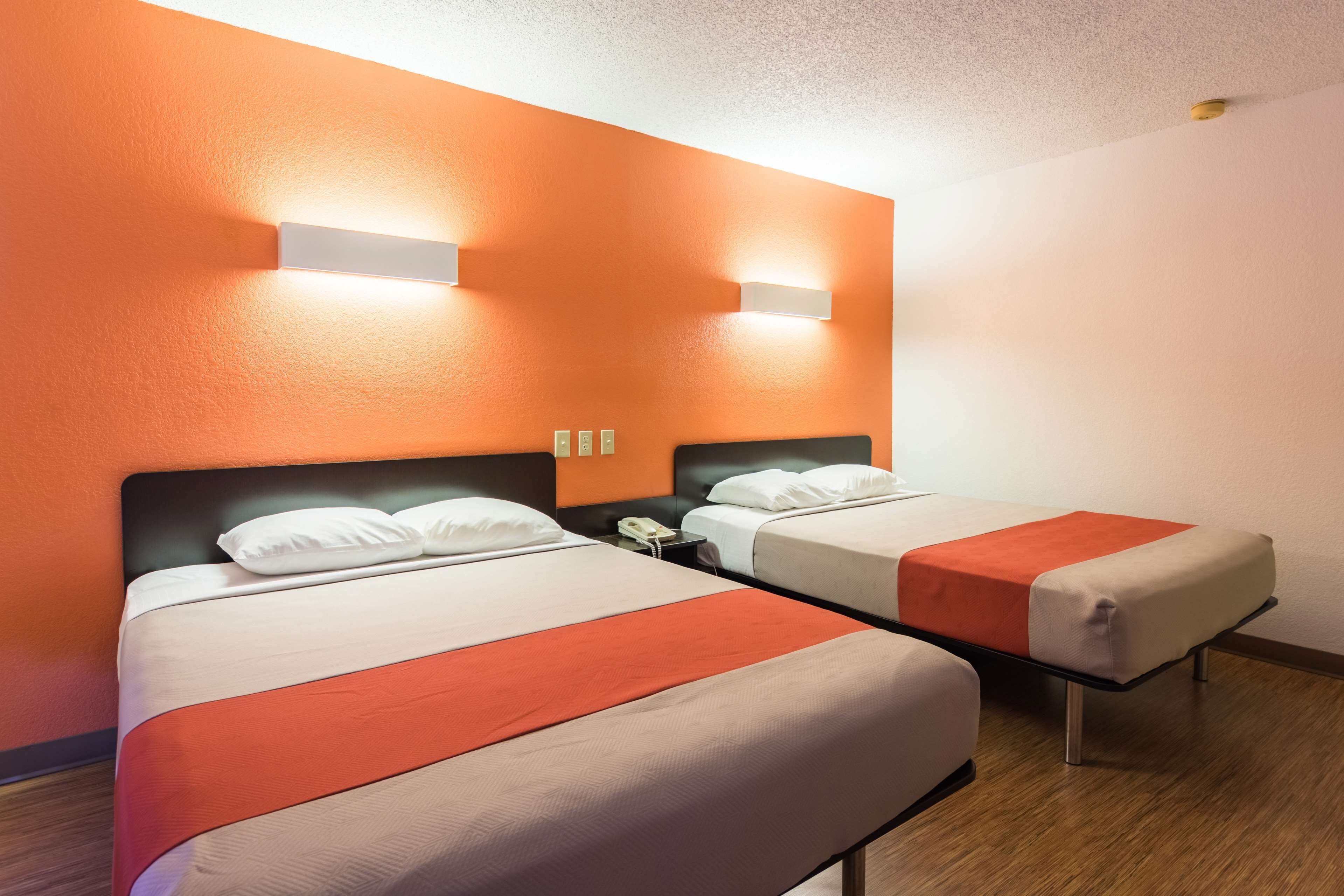 Hotellihuone, jossa on kaksi sänkyä, ja molemmissa on valkoinen ja oranssi sängynpeite. Lisäksi huoneessa on yöpöytä, jossa on puhelin.