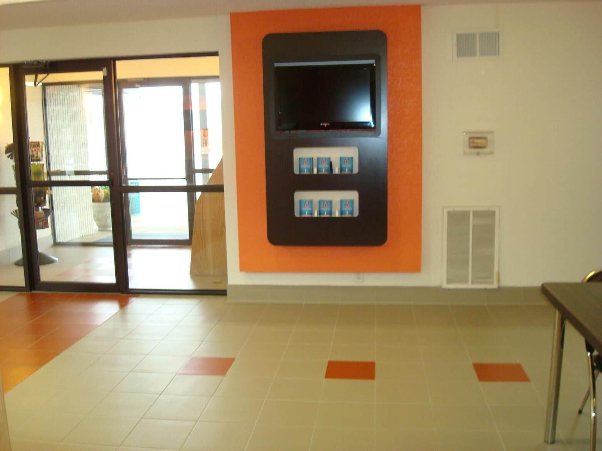 A hotel lobby with a large television mounted on an orange wall, a reception desk, and a tiled floor with orange accents.