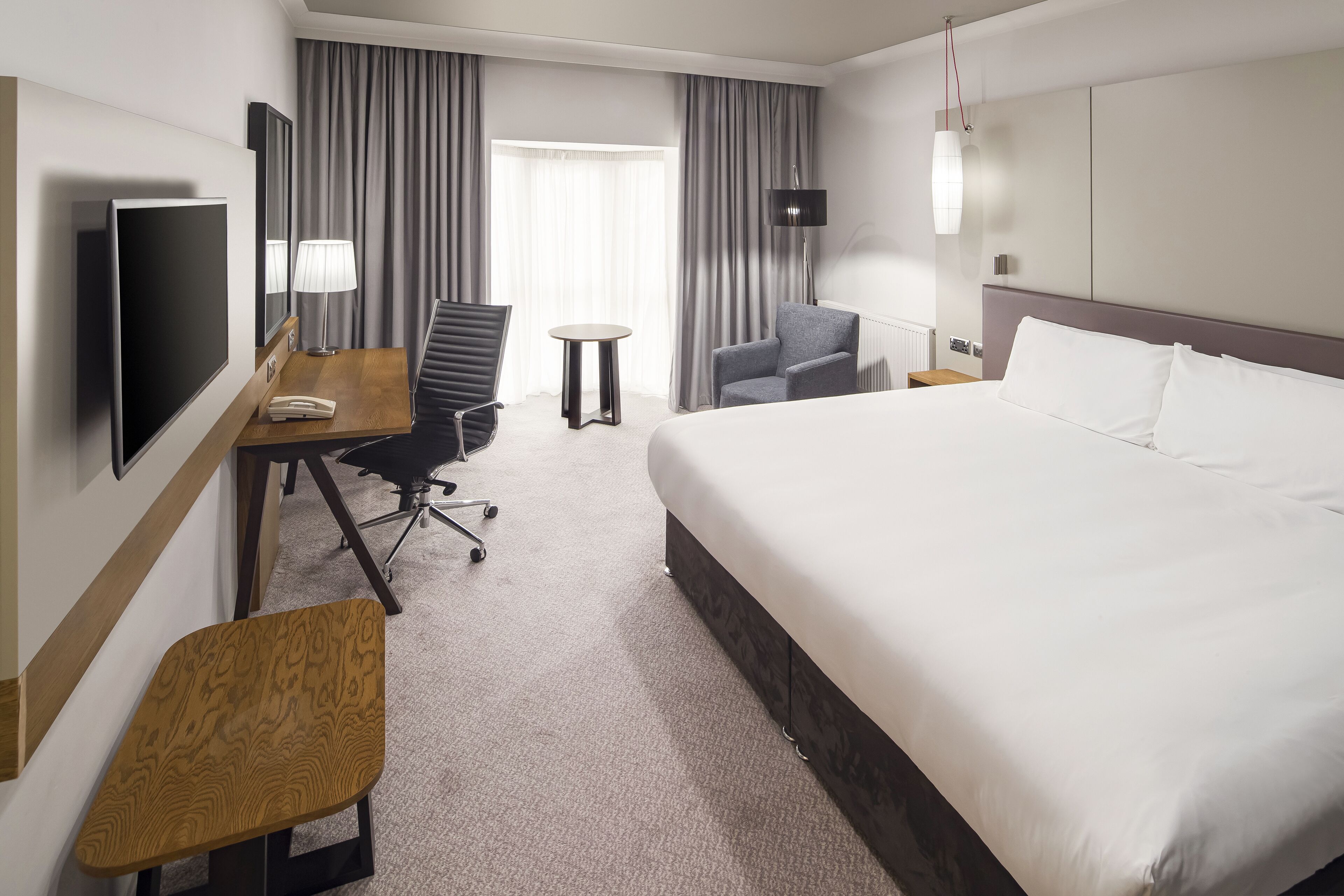 A hotel room with a large bed, a desk with a chair, a TV, a lamp, and a small table.