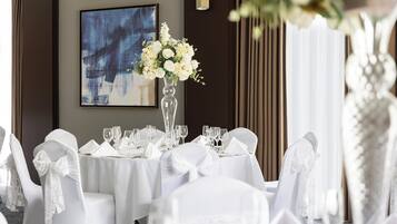 Une salle de banquet avec des tables rondes dressées pour un événement formel, des chaises blanches ornées de nœuds, un vase de fleurs et un tableau abstrait encadré accroché au mur.