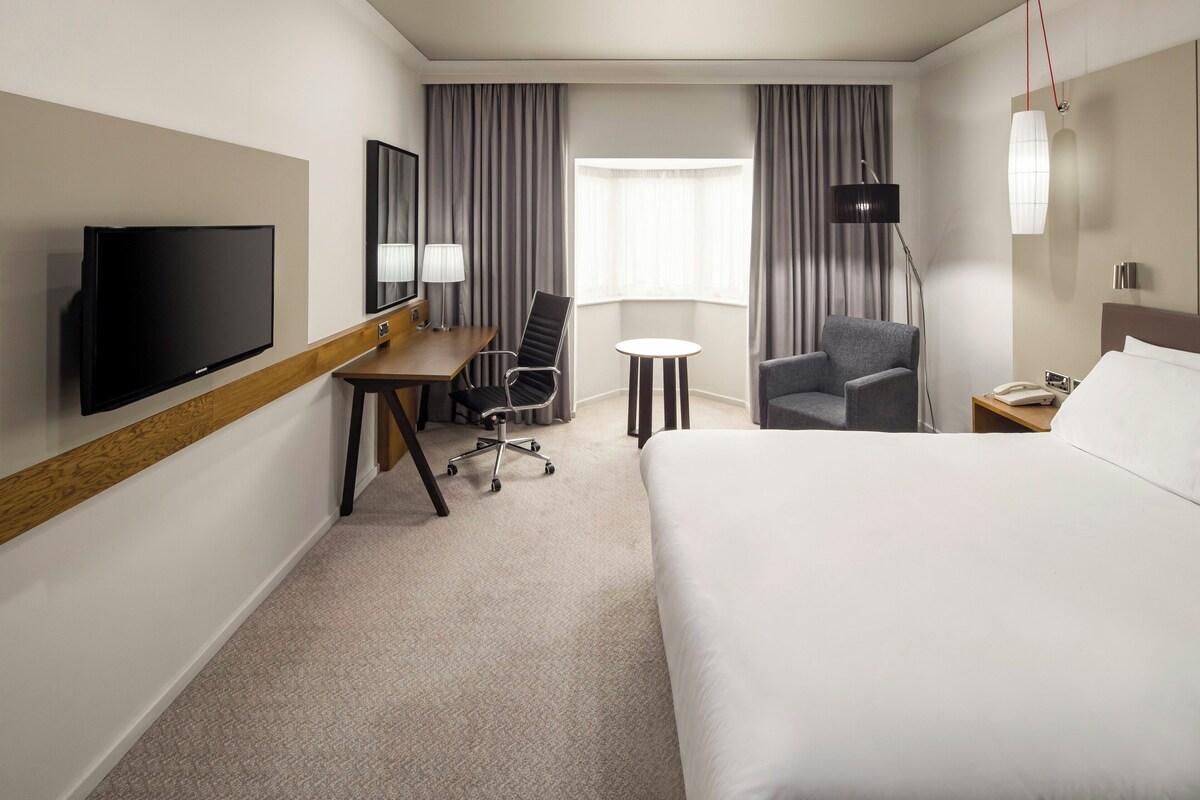 A hotel room with a bed, desk, chair, TV, lamp, and window.