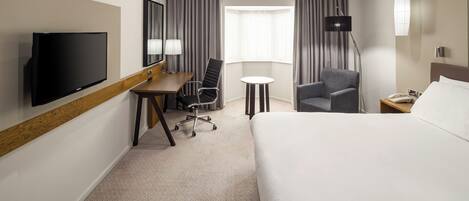 A hotel room with a bed, desk, chair, TV, lamp, and window.