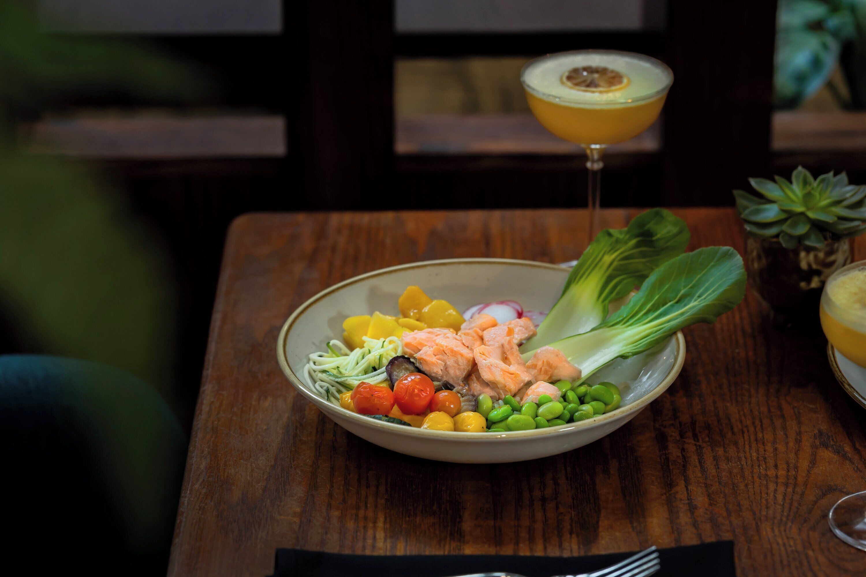A bowl of food with vegetables and salmon on a wooden table.