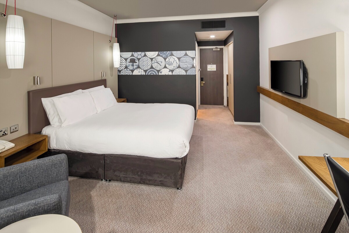 A hotel room with a large bed, a desk, a chair, a television, and a patterned wall.
