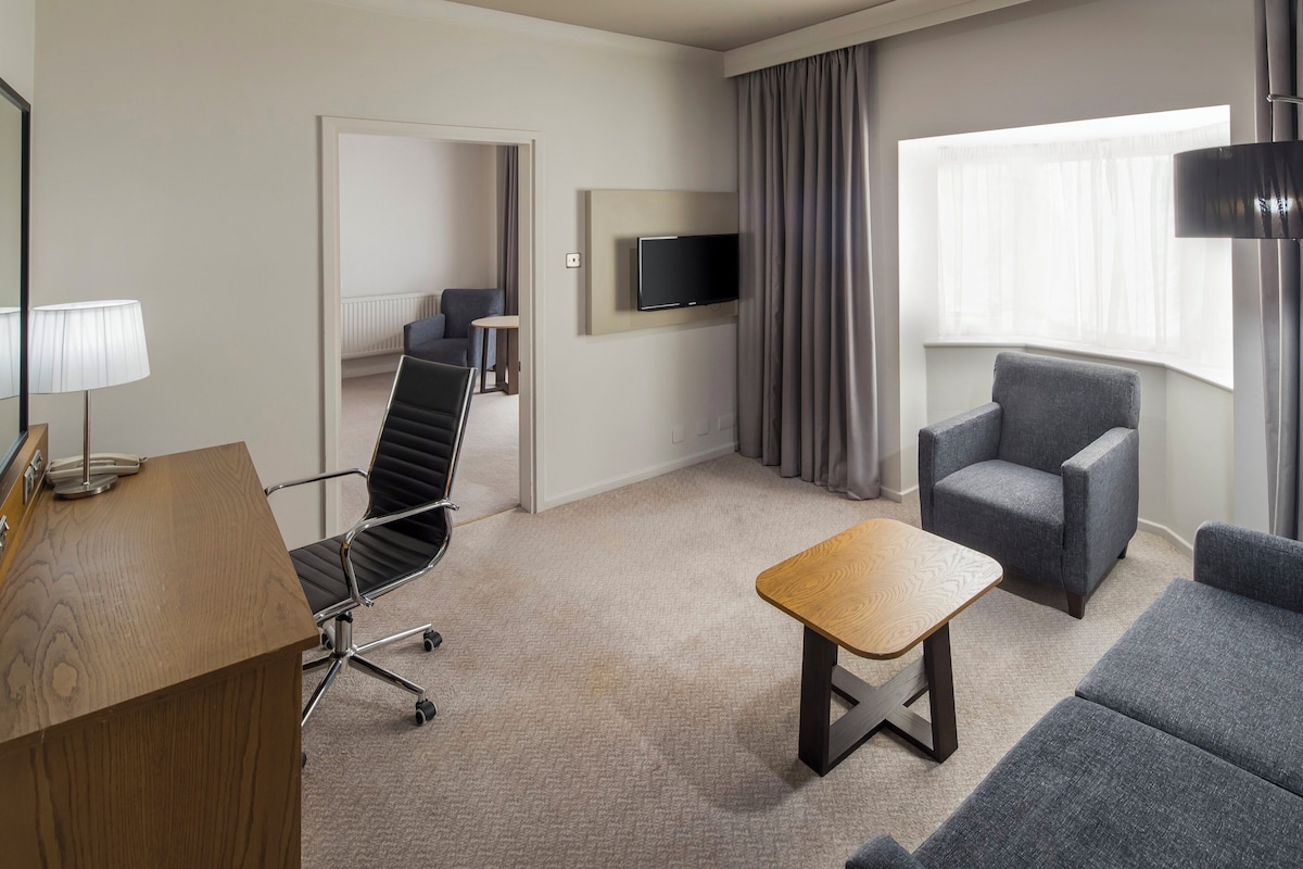 A hotel room with a desk, a chair, a sofa, a TV mounted on the wall, and a window with curtains.