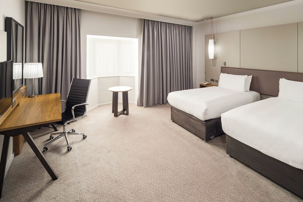 A hotel room with two beds, a desk with a chair, a small round table, and a lamp.