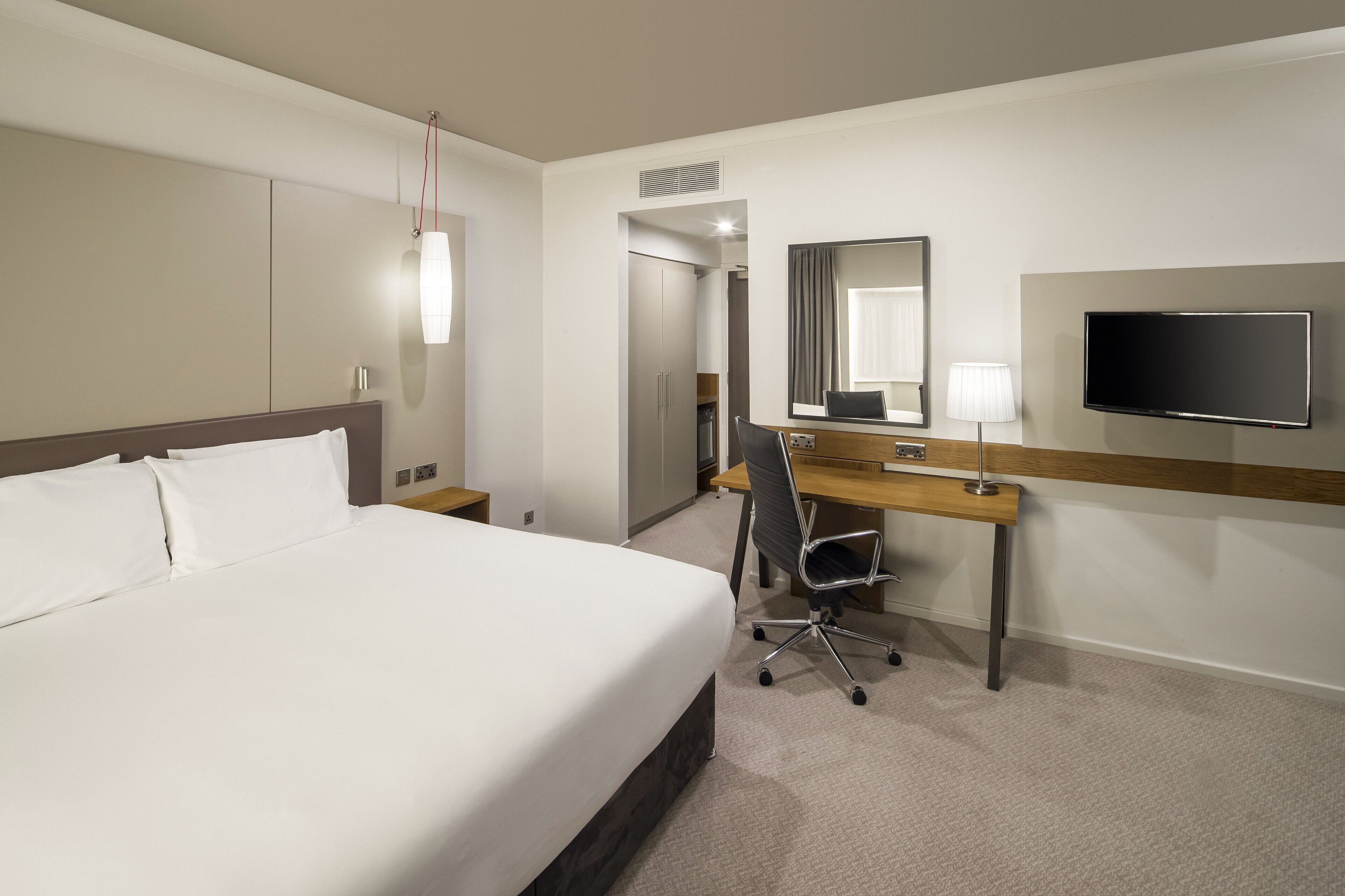 Una habitación de hotel con una cama grande, un escritorio con silla, un televisor y un espejo.