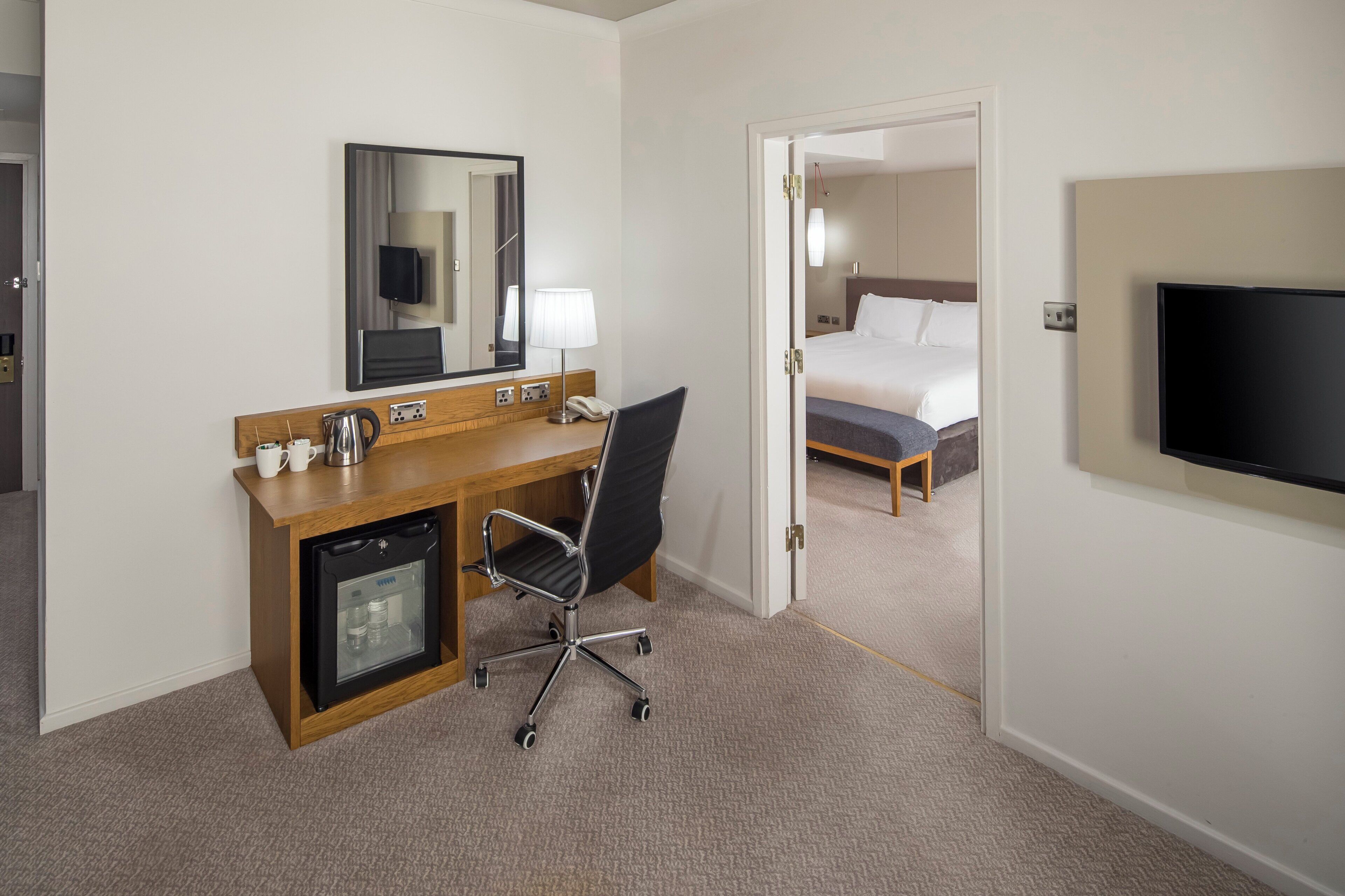 Habitación de hotel con escritorio de madera, una silla, un mini refrigerador, una cama, un televisor y una puerta que da a otra habitación.