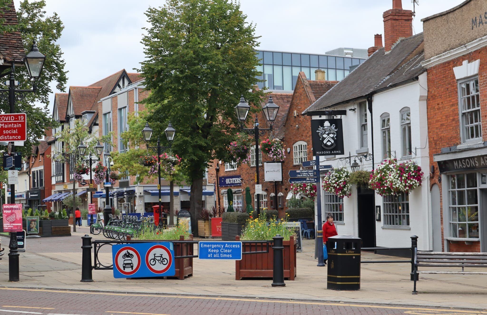 Een straatbeeld met traditionele gebouwen, een bord voor de pub 'Masons Arms', en een waarschuwingsbord met de tekst 'Keep Clear at all times'.